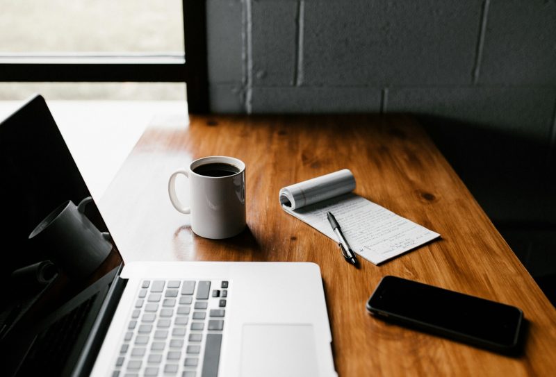 A work desk with laptop, notepad, coffeee cup, cell phone and pen sitting beside each other
