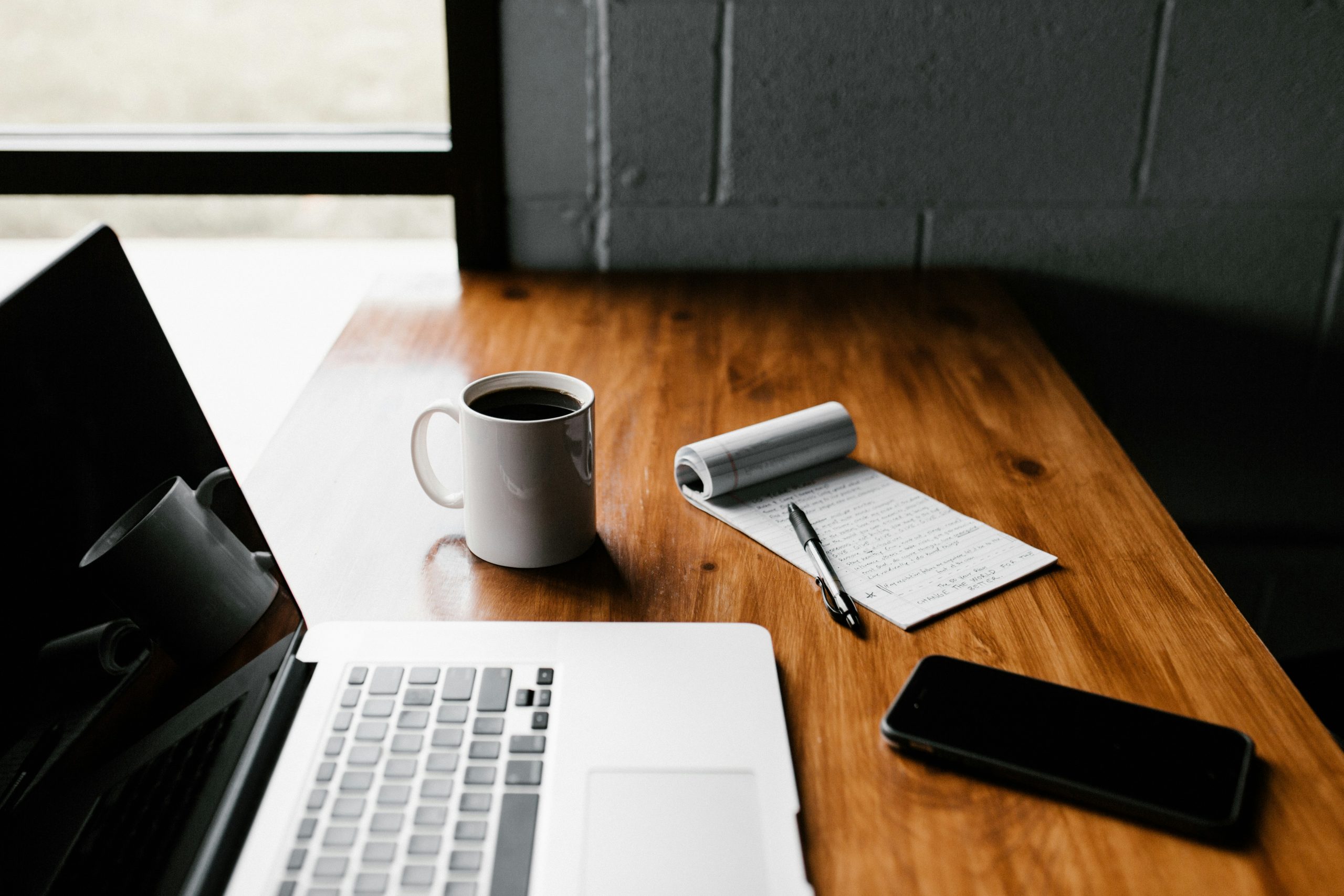 A work desk with laptop, notepad, coffeee cup, cell phone and pen sitting beside each other