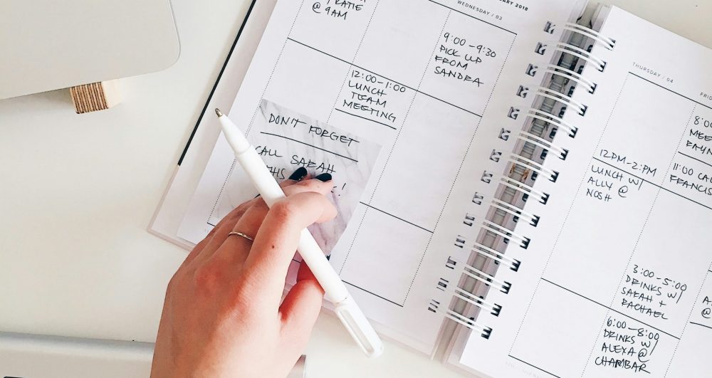 marissa-grootes-flRm0z3MEoA-unsplash An organised work desk from the top view, a hand is holding a pen and turning the page of a calendar note book.