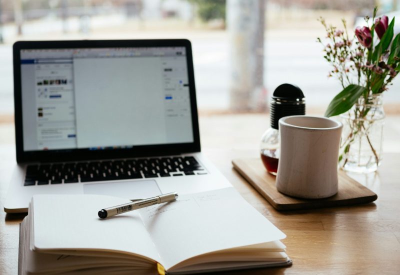 nick-morrison-FHnnjk1Yj7Y-unsplash A home work desk with laptop computer, coffee cup and a notepad with a pen resting on it.
