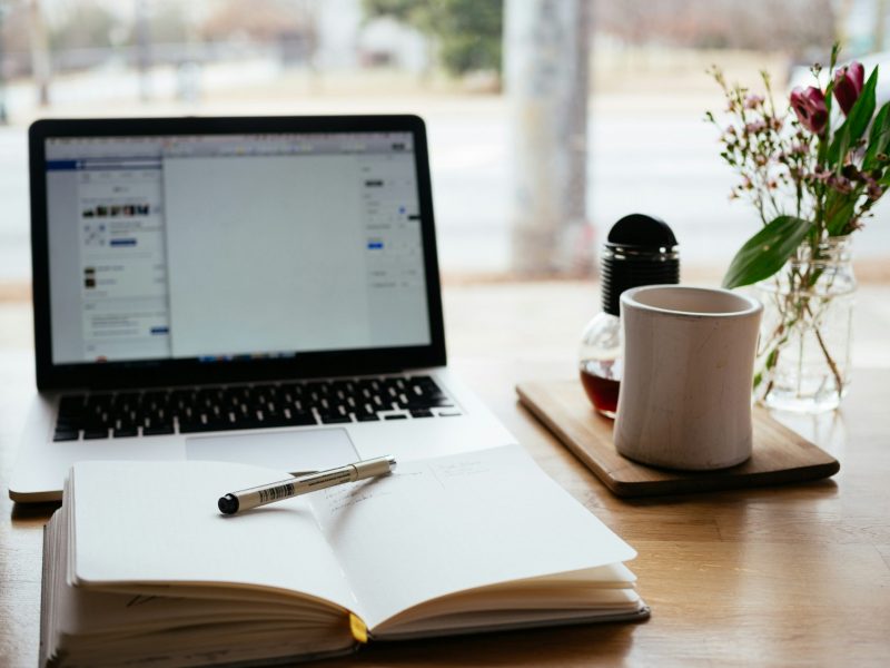 nick-morrison-FHnnjk1Yj7Y-unsplash A home work desk with laptop computer, coffee cup and a notepad with a pen resting on it.